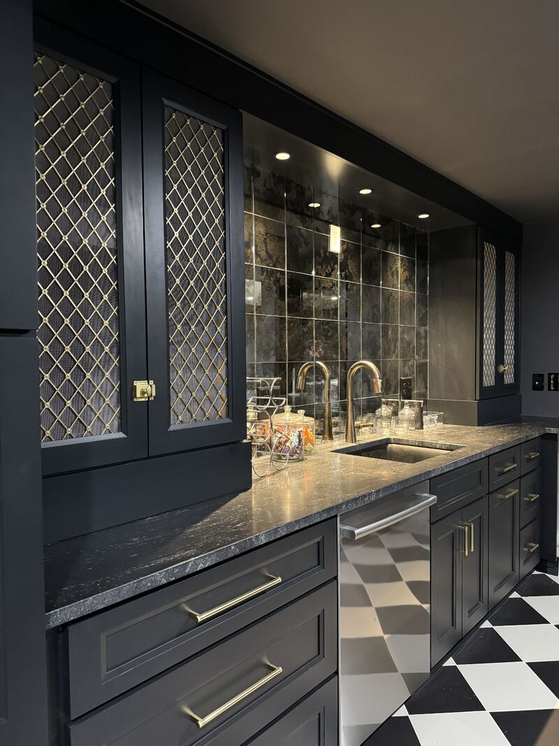 Dramatic basement wet bar with charcoal black shaker cabinets, gold wire mesh cabinet doors, dark granite countertop, antique mirror tile backsplash, brass hardware and faucet, and black and white checkered tile floor 