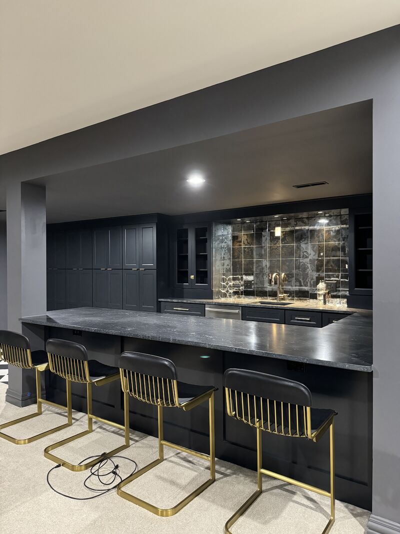 Basement wet bar with charcoal black shaker cabinets, dark granite countertop, antique mirror tile backsplash, and gold bar stools