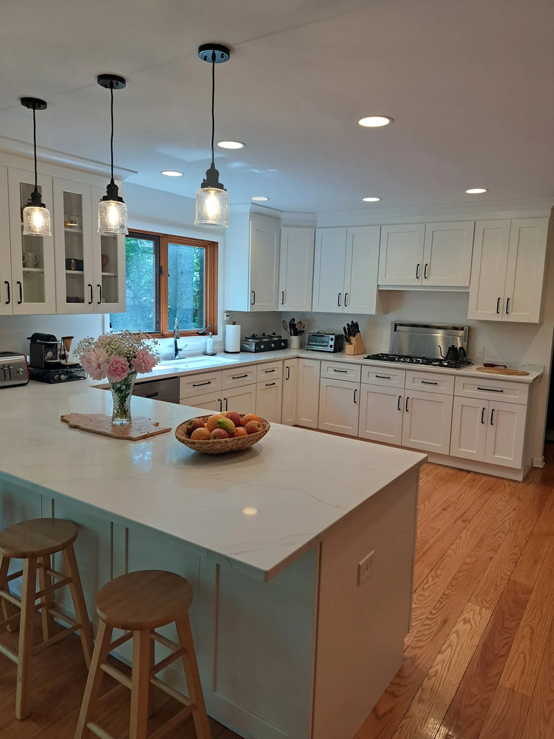 White shaker kitchen with marble quartz island and countertops, glass-front upper cabinets, matte black hardware, seeded glass pendant lights, and warm oak hardwood floors