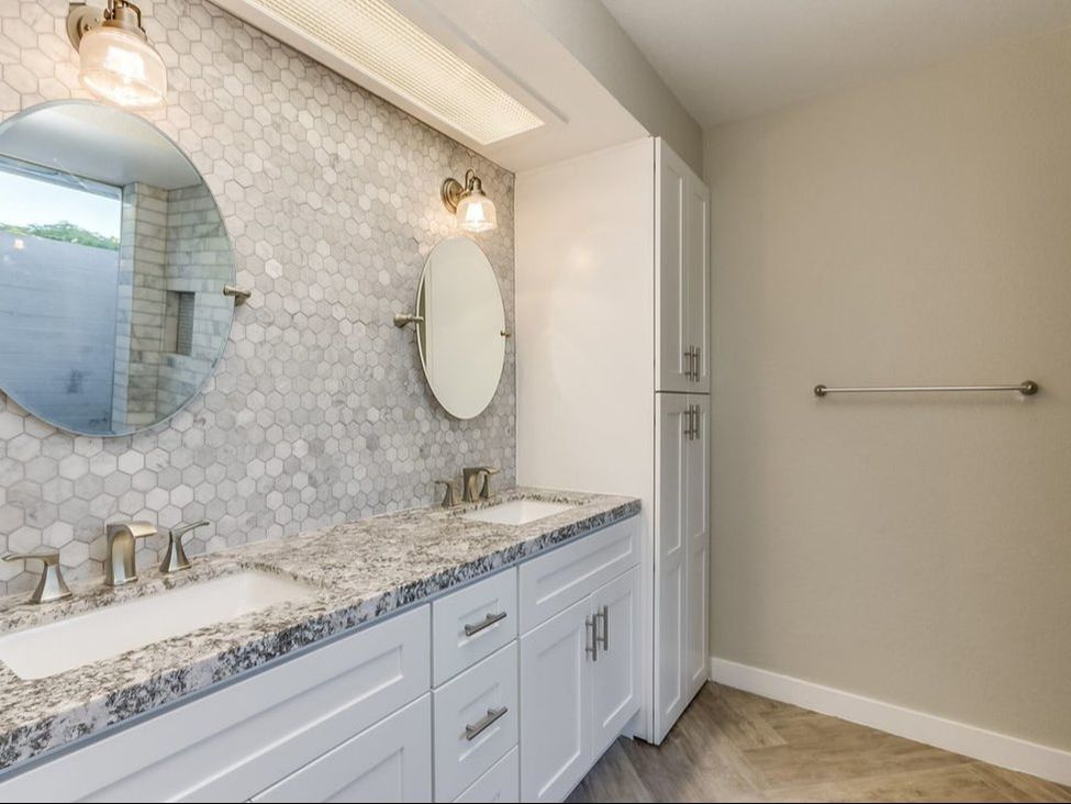 Double sink white shaker vanity with brushed nickel widespread faucets, granite countertop, hexagon tile backsplash, and round mirrors