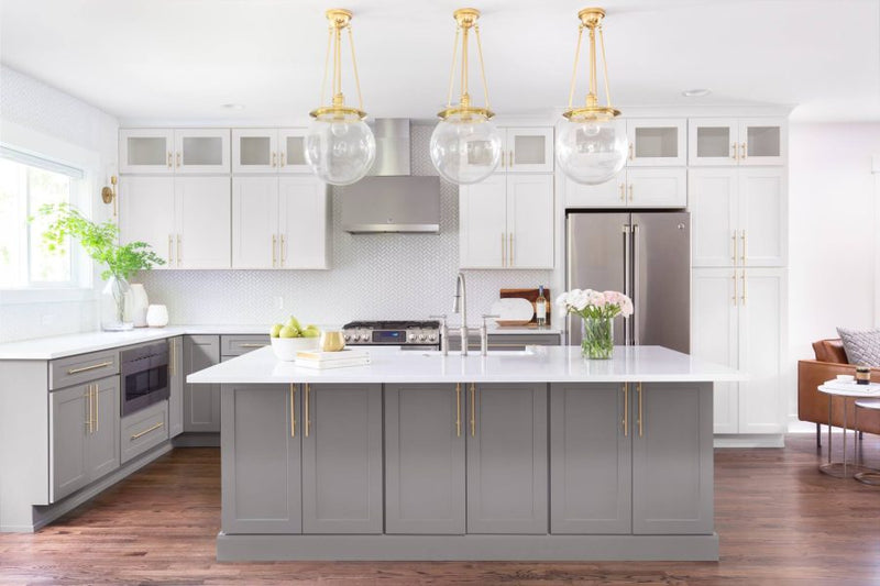 Two-tone kitchen with grey shaker island, white upper cabinets with glass fronts, brass hardware, herringbone tile backsplash, and globe pendant lighting