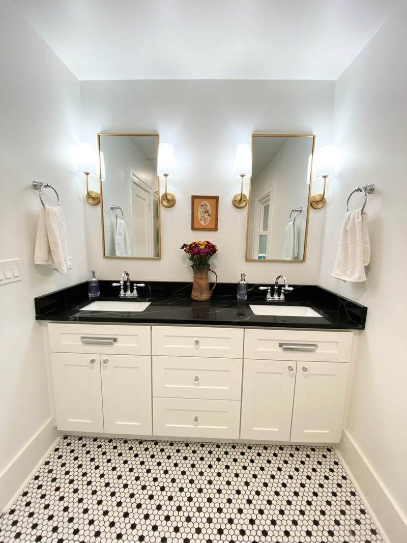 White shaker double vanity with black granite countertop, dual undermount sinks, brushed nickel hardware, gold framed mirrors, brass sconces, and black and white mosaic tile floor