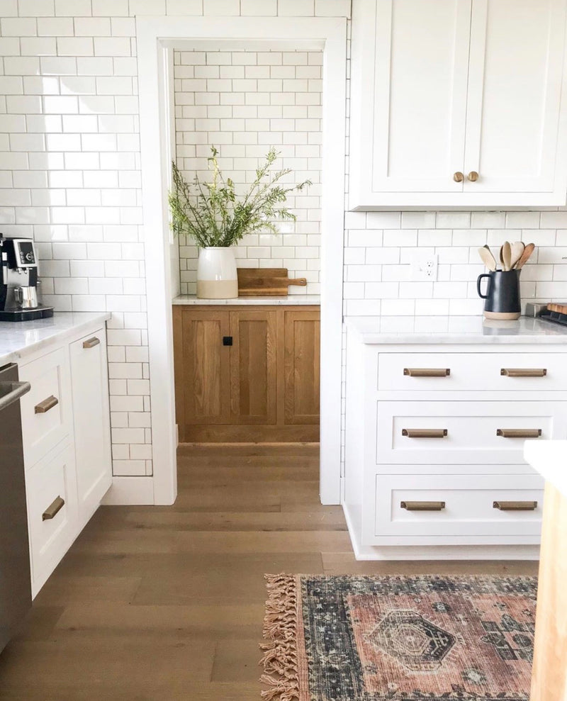 White shaker kitchen with marble countertop, gold hardware, white subway tile backsplash, natural wood pantry cabinet, and wide plank hardwood floors