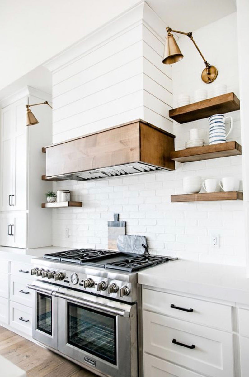 White shaker kitchen cabinets with shiplap range hood, wood floating shelves, subway tile backsplash, and brass sconce lighting