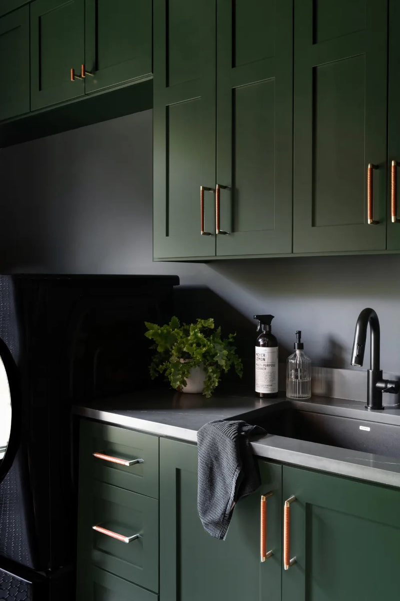 Dark green shaker cabinets with leather-wrapped copper cabinet pulls, black countertop, and matte black faucet in a moody laundry room