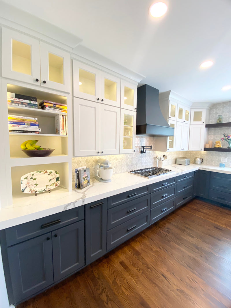 Two-tone kitchen with navy blue base cabinets, white upper cabinets with glass fronts, open shelving, quartz countertops, and patterned tile backsplash