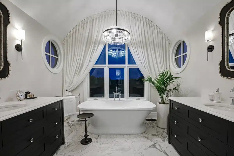 Luxury primary bathroom with dual black vanities, freestanding soaking tub, marble tile floors, crystal chandelier, and vaulted arch ceiling
