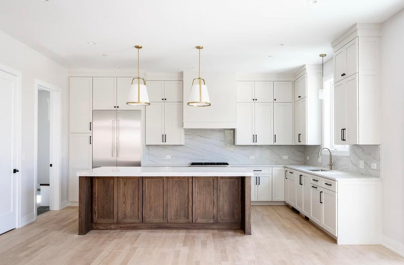 Bright white shaker kitchen with stained wood panel island, full-height marble slab backsplash, white quartz countertop, black hardware, gold pendant lights, and light hardwood floors