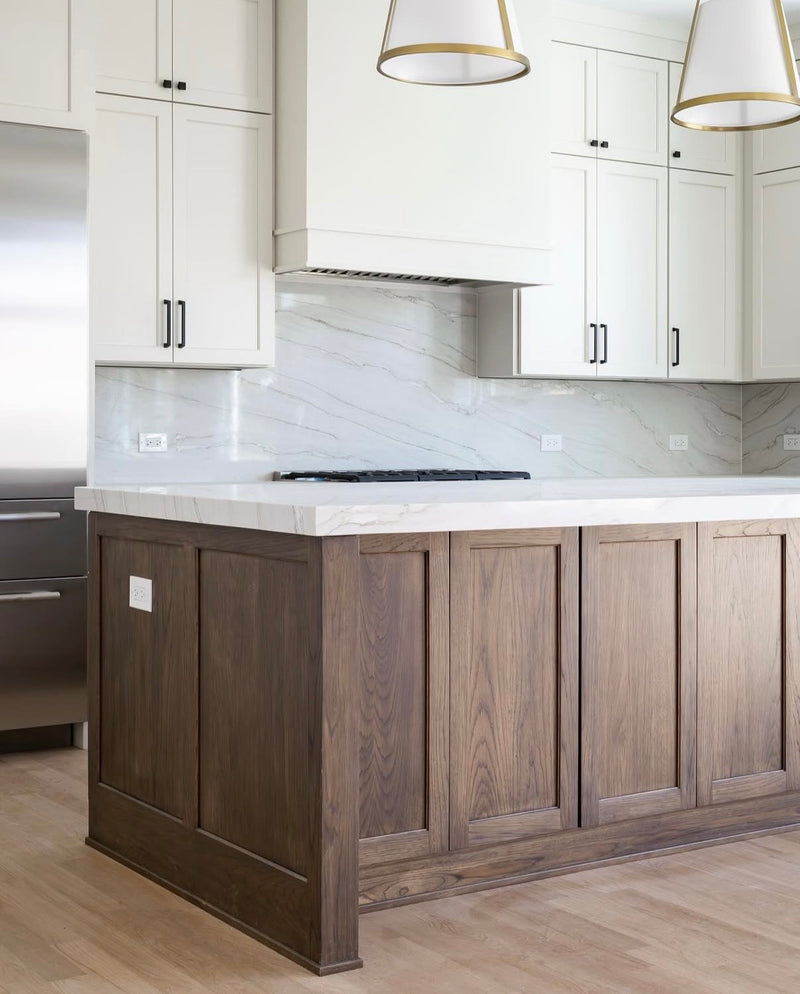 Two-tone kitchen with white shaker upper cabinets, stained wood island with panel detail, marble slab backsplash, white quartz countertop, and gold drum pendant lights