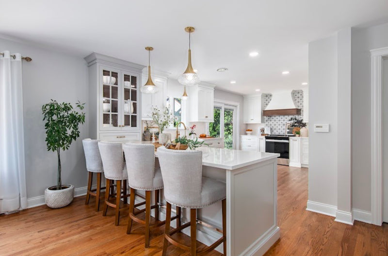 White shaker kitchen with grey island, glass front cabinet hutch, brass pendant lighting, upholstered bar stools, and wood range hood