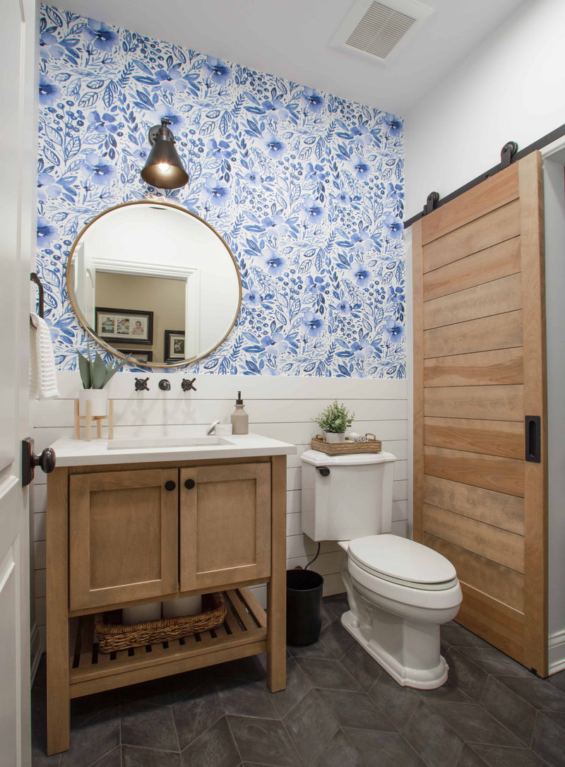 Farmhouse bathroom with natural wood vanity, white quartz countertop, blue floral wallpaper accent wall, shiplap wainscoting, wood barn door, gold round mirror, and dark hex tile floor