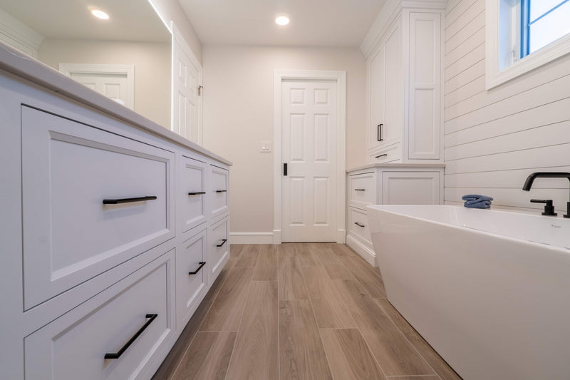 Primary bathroom with white shaker vanity drawers, matte black hardware, freestanding soaking tub, shiplap accent wall, and wood-look tile floor