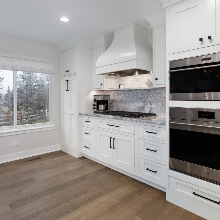 White shaker kitchen with gray marble countertop and backsplash, curved white range hood, built-in double wall ovens, matte black hardware, and medium hardwood floors
