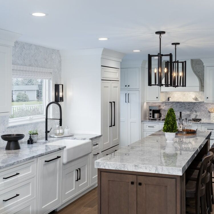 Two-tone kitchen with white shaker cabinets, dark wood island, marble countertops, farmhouse sink, black hardware, and stone mosaic backsplash 