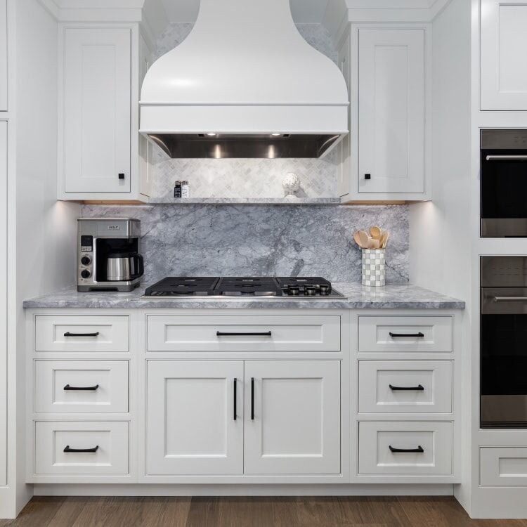 White shaker kitchen with gray marble countertop and full-height slab backsplash, curved white range hood, matte black hardware, gas cooktop, and double wall ovens