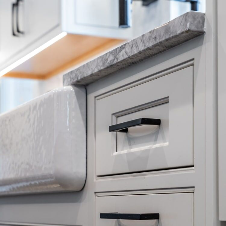 Close-up of matte black square cabinet pulls on white shaker drawer fronts with marble countertop and farmhouse sink