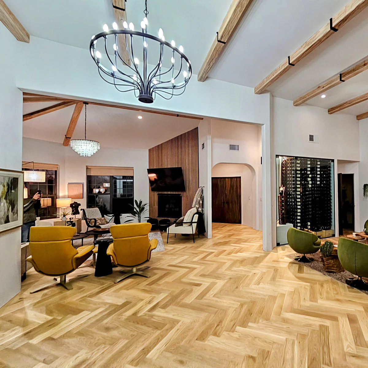 Open-concept living room with natural oak herringbone hardwood flooring, vaulted ceiling with wood beams, and modern chandelier