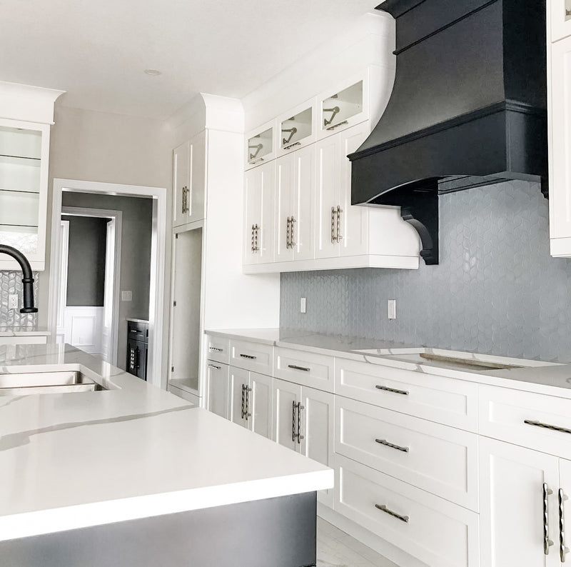 White shaker kitchen with black statement range hood, blue hexagon tile backsplash, glass-front upper cabinets, white quartz countertop, and brushed nickel hardware