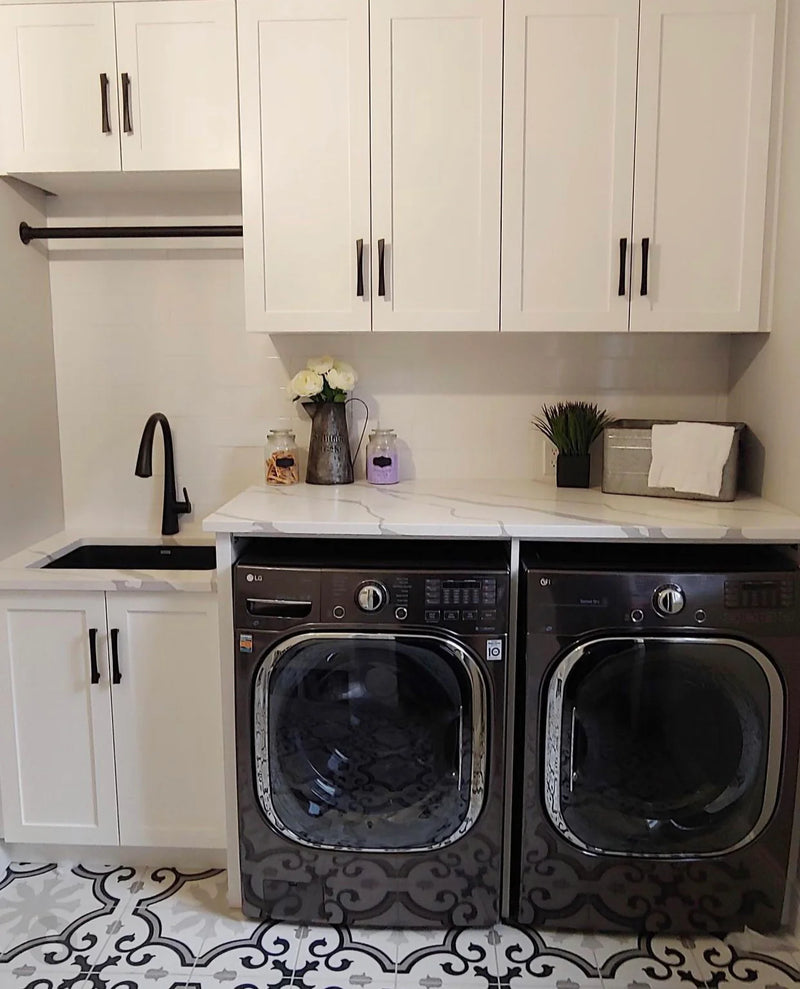 Laundry room with white shaker cabinets, marble quartz countertop, matte black undermount sink and faucet, front-load washer and dryer, and patterned encaustic tile floor