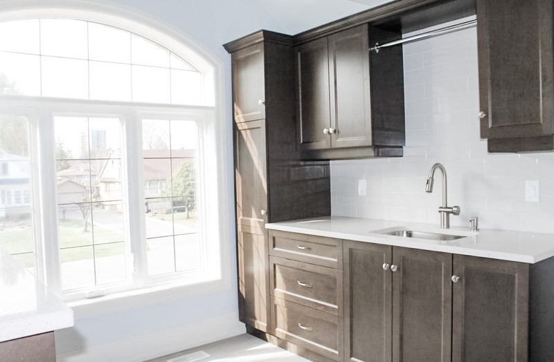Laundry room with dark stained shaker cabinets, tall pantry cabinet, white quartz countertop, undermount sink, brushed nickel faucet, built-in hanging rod, and arched window