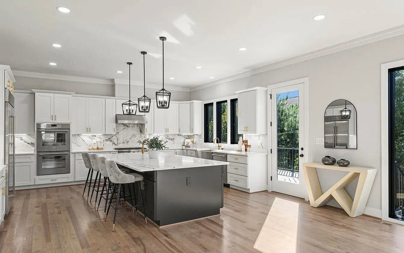 White shaker kitchen cabinets with black island, marble slab backsplash, brass hardware, cage pendant lighting, and hardwood floors