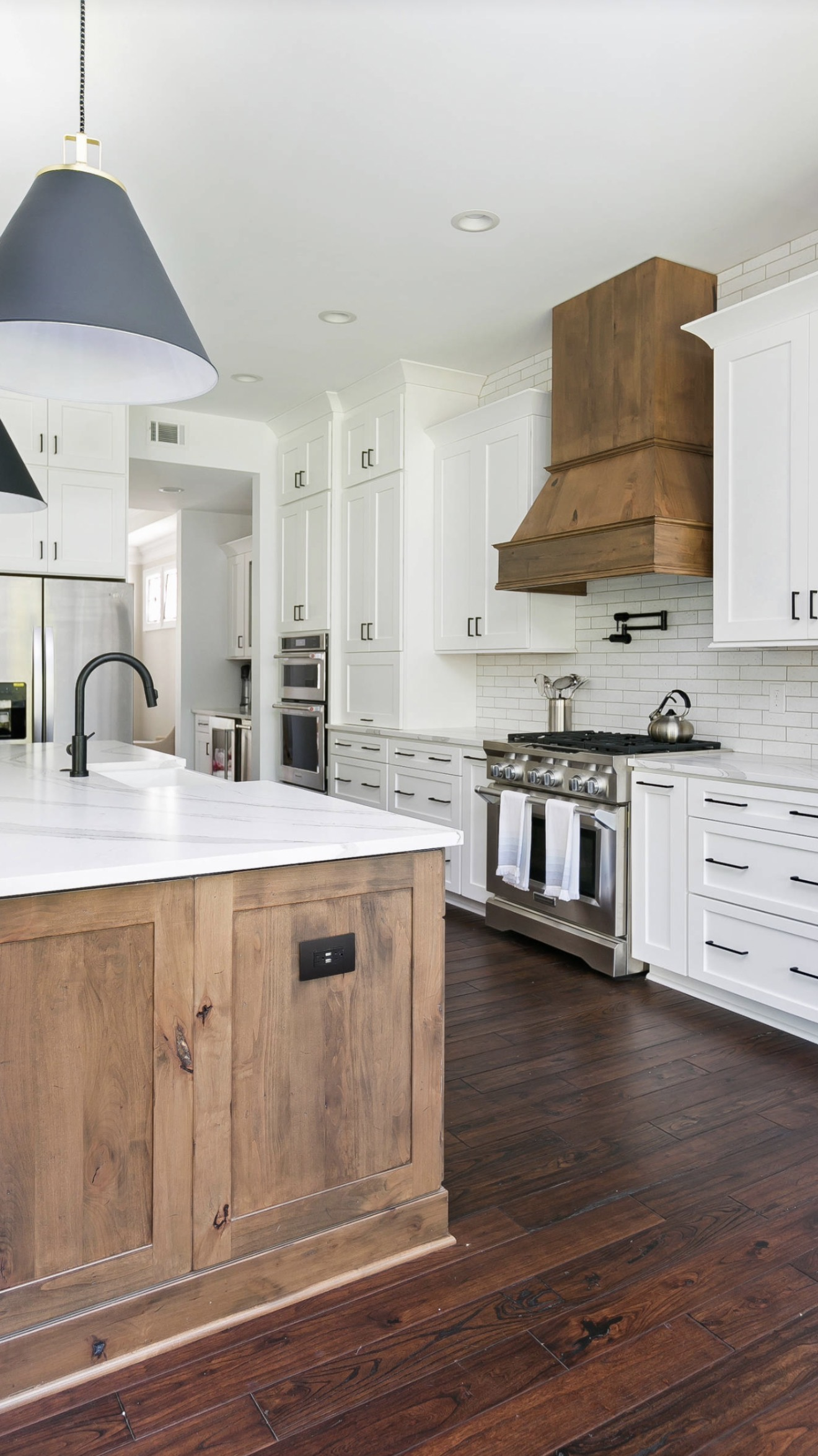 White kitchen featuring quartz countertops, a wood island base, custom wood range hood, matte black hardware, and pendant lighting, highlighting layered kitchen finishes and natural wood accents