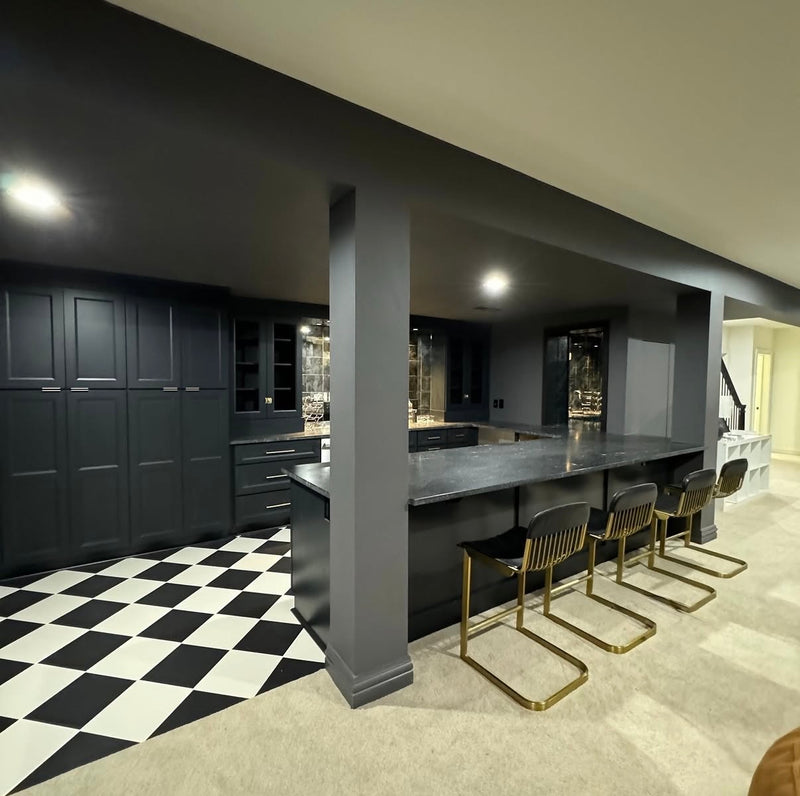 Dramatic basement bar with charcoal black shaker cabinetry, dark granite countertop, gold bar stools, black and white checkered tile floor, and antique mirror backsplash