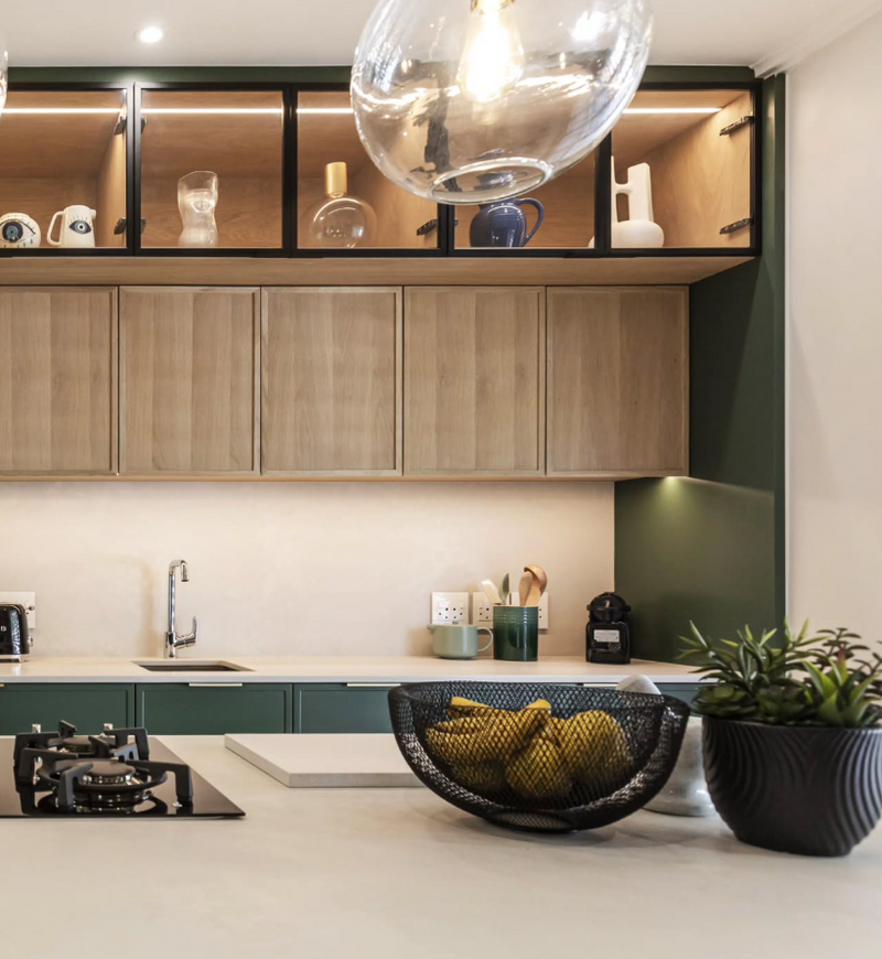 Modern kitchen with deep green lower cabinets, white oak upper cabinets with open display shelving and under-cabinet lighting, cream countertop, and globe pendant light