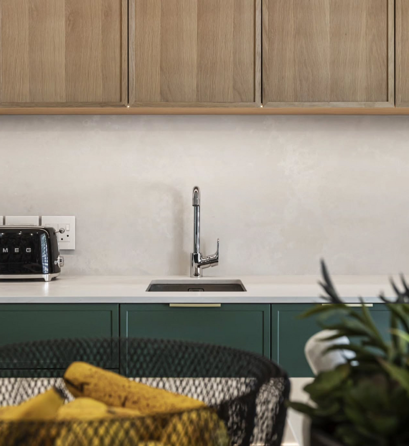 Two-tone kitchen detail with deep green lower cabinets, white oak upper cabinets, cream countertop, undermount sink, and plaster finish backsplash