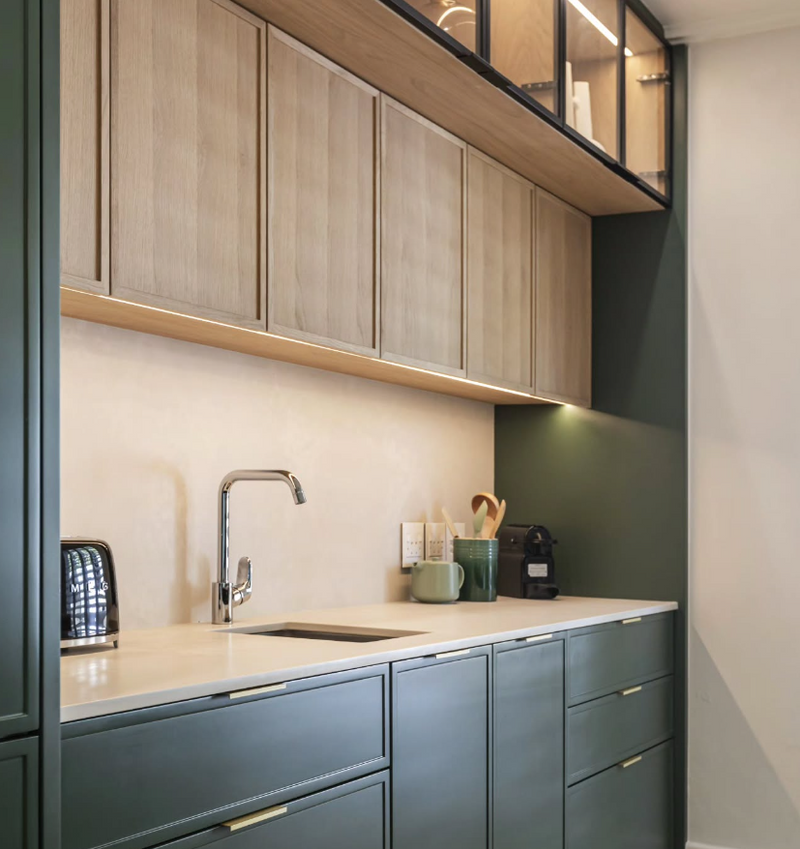 Two-tone kitchen with deep green lower cabinets, white oak upper cabinets with black frame glass doors, under-cabinet lighting, cream countertop, and undermount sink
