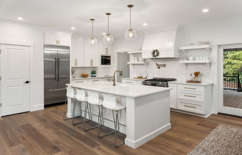 All-white kitchen with shaker cabinets, large center island, white quartz countertop, floating shelves, white range hood, gold hardware, glass globe pendant lights, and dark hardwood floors