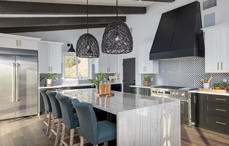 Two-tone kitchen with white upper shaker cabinets, dark lower cabinets, marble waterfall island, geometric tile backsplash, black statement range hood, woven pendant lights, and exposed dark ceiling beams