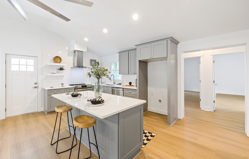 Gray shaker kitchen with matching island, white quartz countertop, white brick backsplash, open floating shelves, stainless steel range hood, and light hardwood floors
