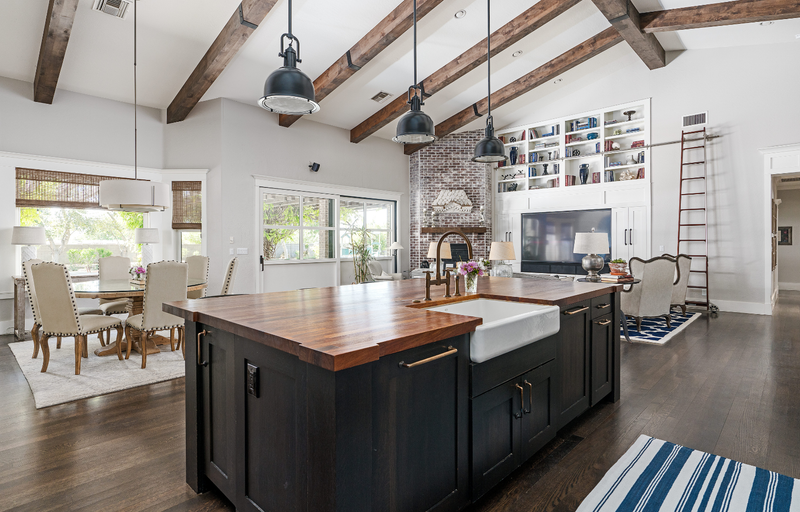 Open concept farmhouse kitchen with black shaker island, butcher block countertop, farmhouse sink, bronze faucet and hardware, exposed wood beam vaulted ceiling, and industrial pendant lights