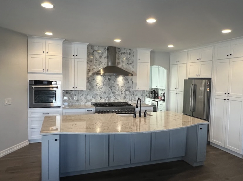 Large kitchen with white shaker cabinets, curved light blue island, gold-veined granite countertop, dramatic floral marble backsplash, stainless steel range hood, and dark hardwood floors
