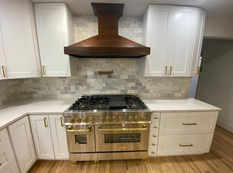 White shaker kitchen cabinets with brass hardware, wood range hood, marble tile backsplash, and professional dual-fuel range