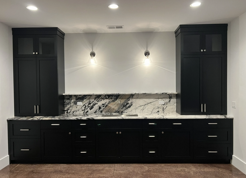 Basement wet bar with floor-to-ceiling black shaker cabinets, dramatic marble slab countertop and backsplash, glass-front upper cabinets, and brushed nickel hardware