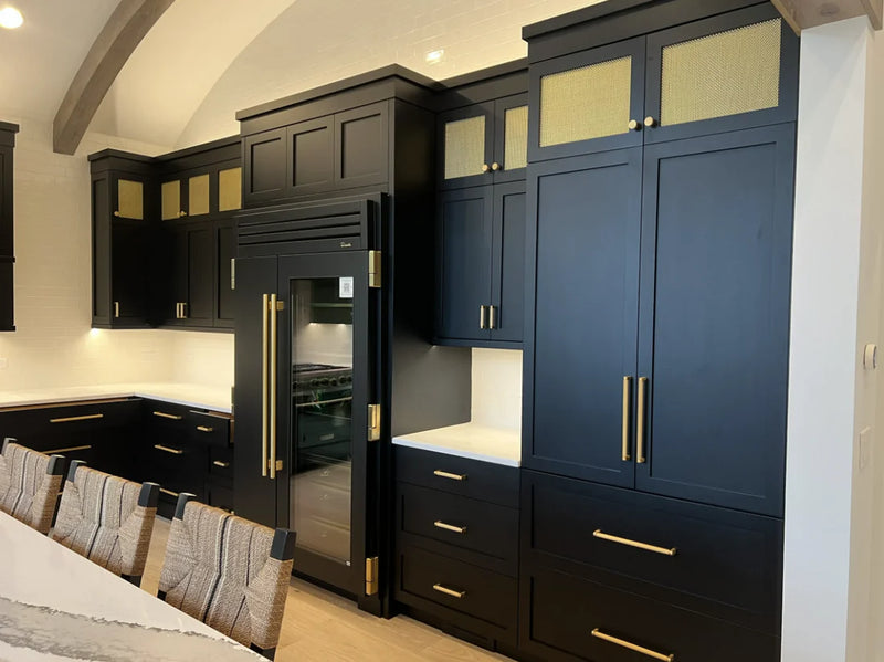 Bold kitchen with floor-to-ceiling black and navy shaker cabinets, gold wire mesh upper cabinet inserts, brass hardware, built-in professional refrigerator, white quartz countertop, and vaulted beam ceiling