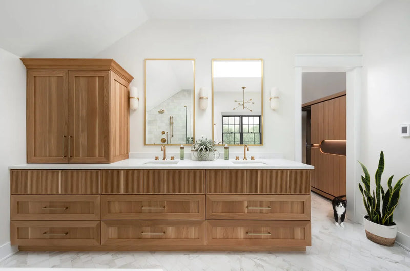 Primary bathroom with natural wood double vanity, fluted drawer fronts, white quartz countertop, gold hardware and faucets, gold framed mirrors, and marble tile floor