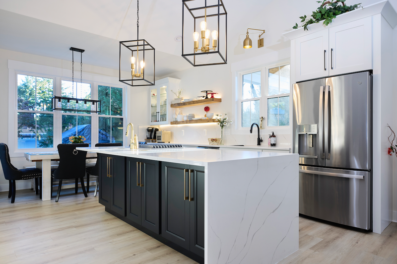 Two-tone kitchen with white shaker cabinets, charcoal black island, white marble quartz countertop, floating wood shelves, gold hardware, black and gold cage pendant lights, and light hardwood floors