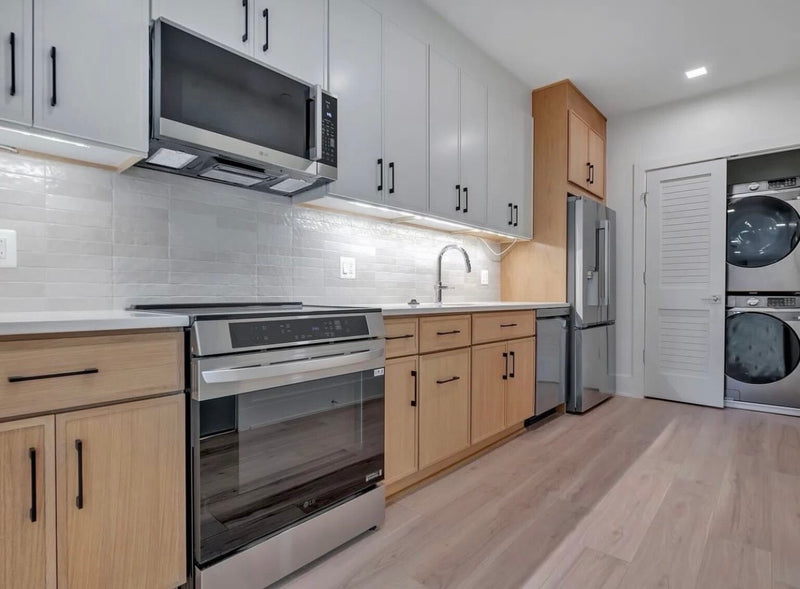 Galley kitchen with two-tone gray upper and natural wood lower cabinets, white quartz countertop, subway tile backsplash, and stacked washer dryer