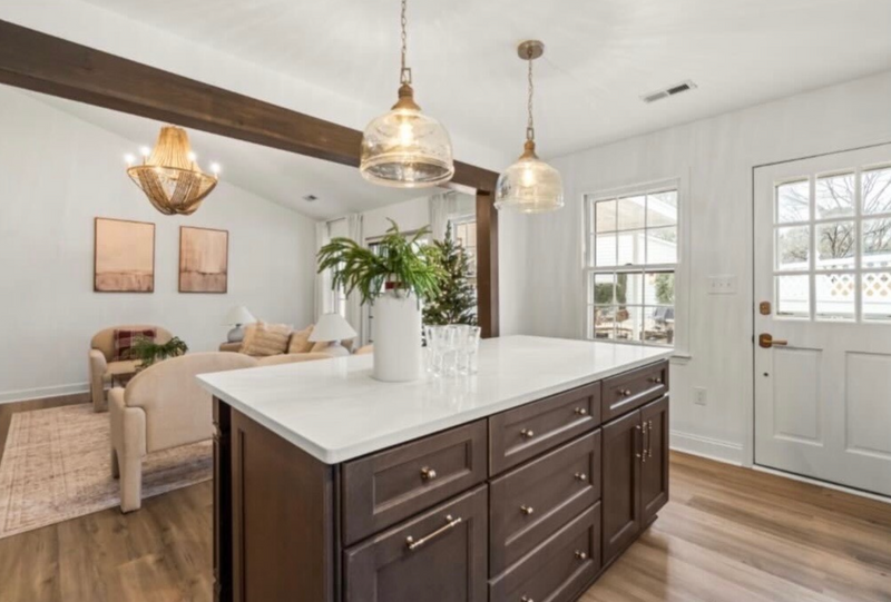 Open concept kitchen with dark brown shaker island, white quartz countertop, gold hardware, glass pendant lights, exposed wood ceiling beam, and light hardwood floors