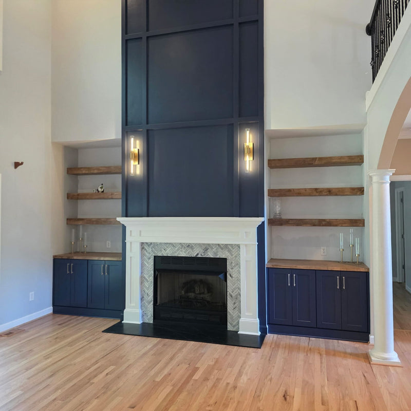 Two-story living room fireplace wall with navy blue board and batten surround, white mantel, herringbone marble tile, flanking navy shaker base cabinets, floating wood shelves, and gold sconces