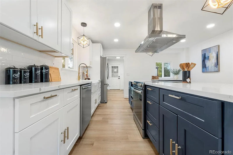 Two-tone kitchen with white shaker cabinets, navy blue island, brass hardware, white quartz countertops, and stainless steel range hood