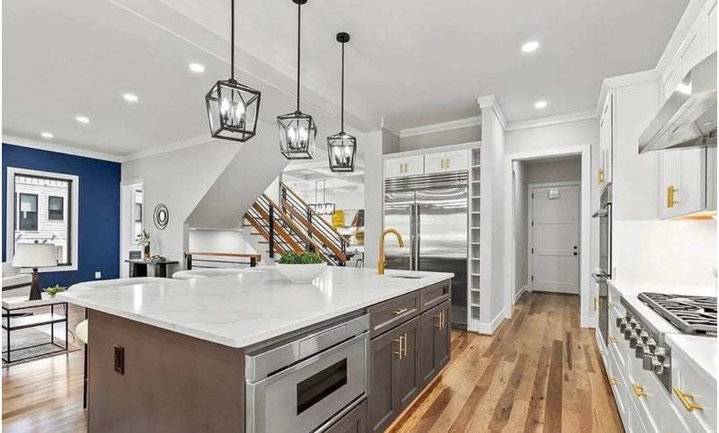 Open concept kitchen with white shaker cabinets, gray island, white quartz countertop, gold hardware and faucet, black lantern pendant lights, built-in microwave drawer, and hardwood floors 